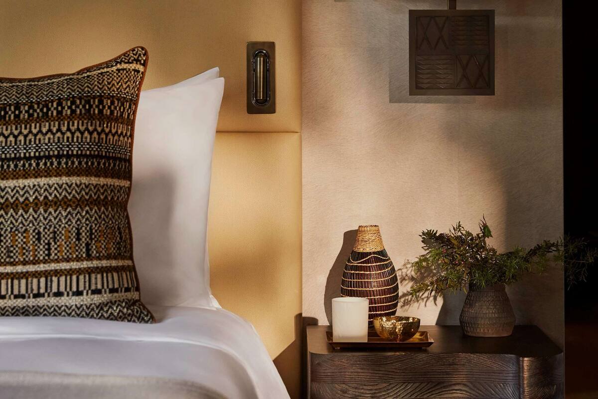 A neatly arranged bedroom with a bed, patterned pillows, a bedside table with a lamp, a vase, and a potted plant.