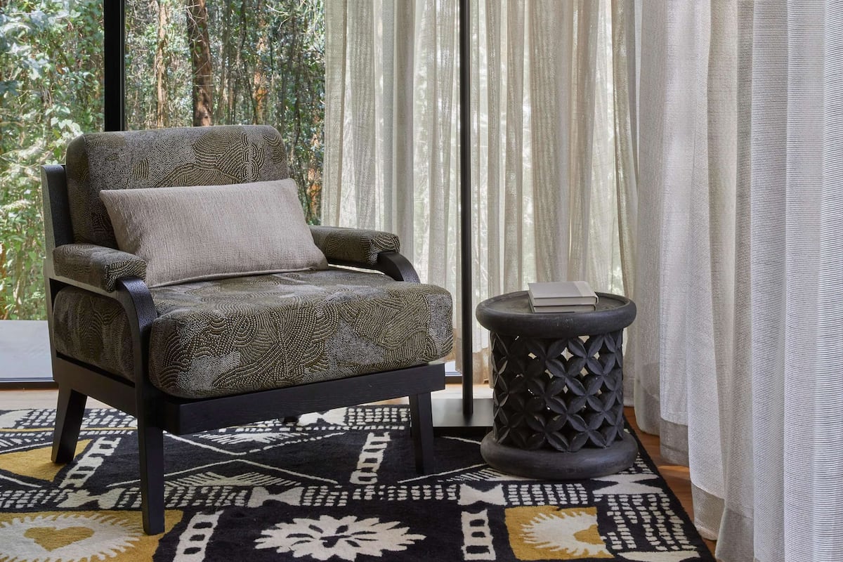 A room with a view of a forest, a patterned armchair, a floor lamp, and a patterned rug.