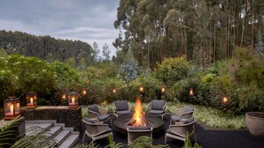 Un espacio de estar al aire libre circular con una chimenea rodeada de sillas de mimbre, frente a un fondo de vegetación exuberante y árboles altos.
