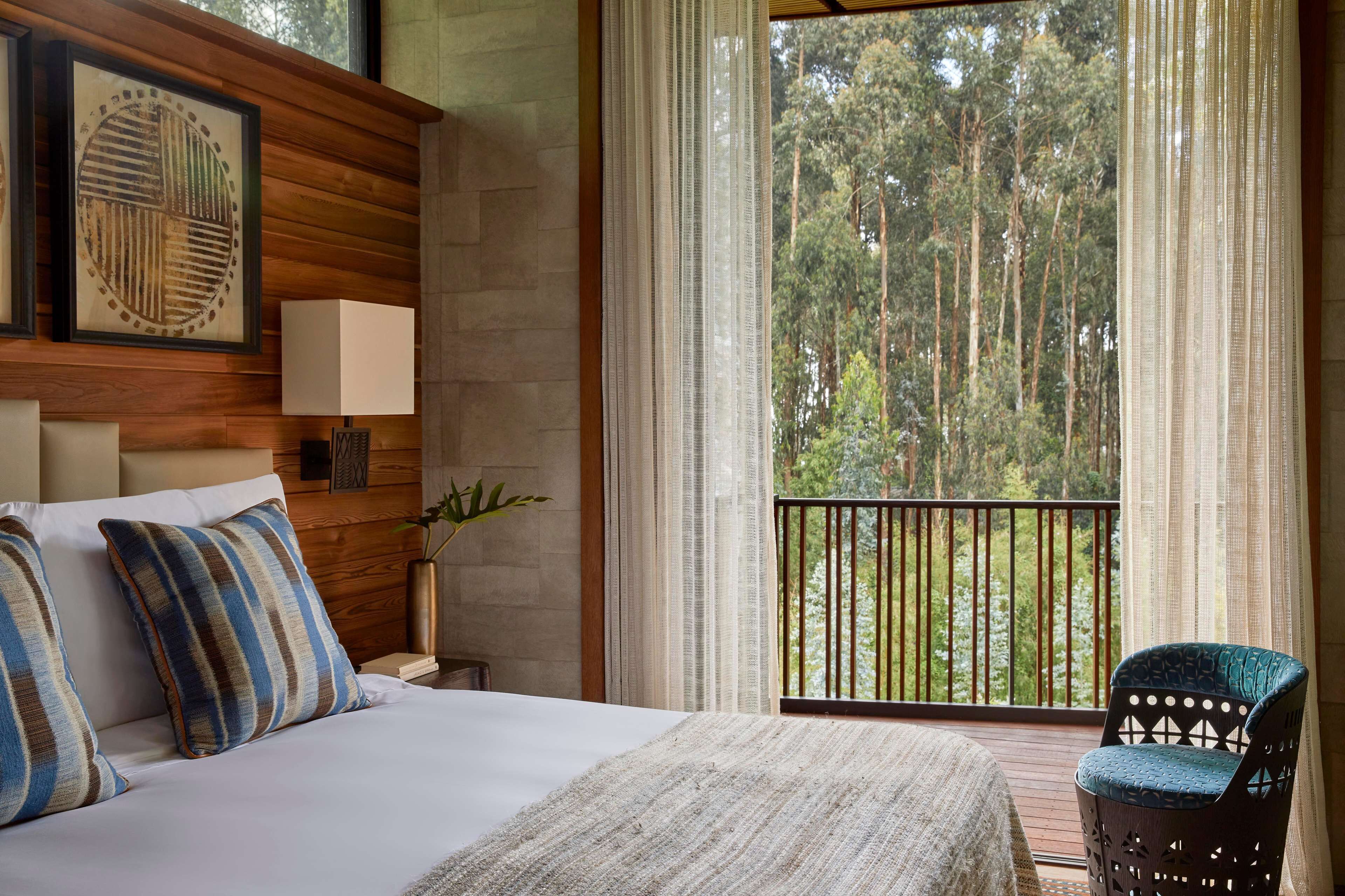 Un dormitorio con cama, almohadas, mesita de noche con lámpara, ventana con cortinas y vista a árboles.