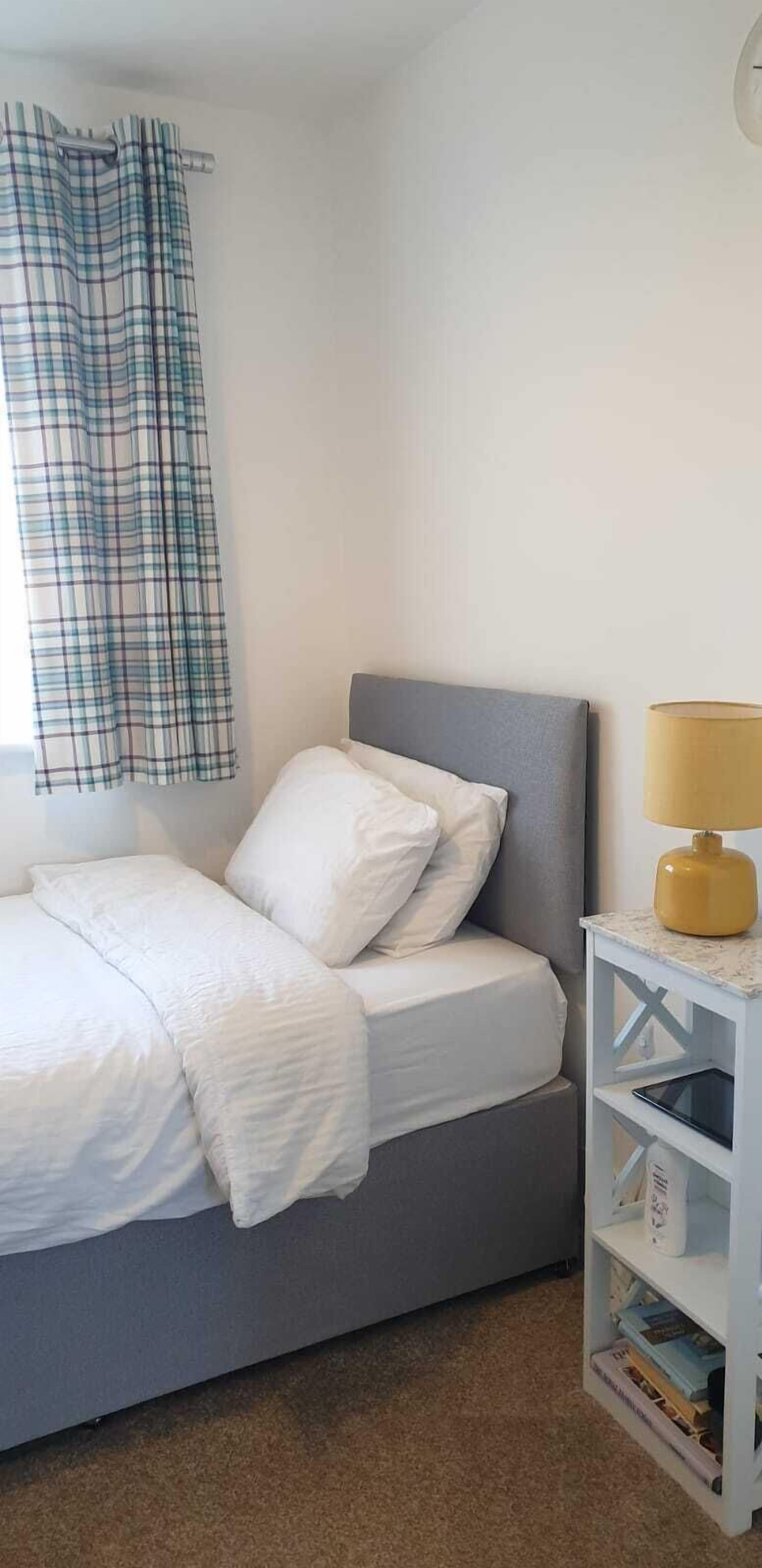 A neatly made bed with white bedding, a grey headboard, and a bedside table with a lamp and books.