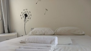 Una cama bien hecha con sábanas y almohadas blancas, una decoración en la pared de un diente de león, y una ventana con cortinas.