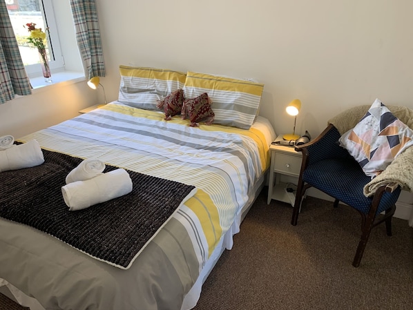 A neatly made bed with striped bedding, a bedside table with a lamp, and a chair with a blanket.