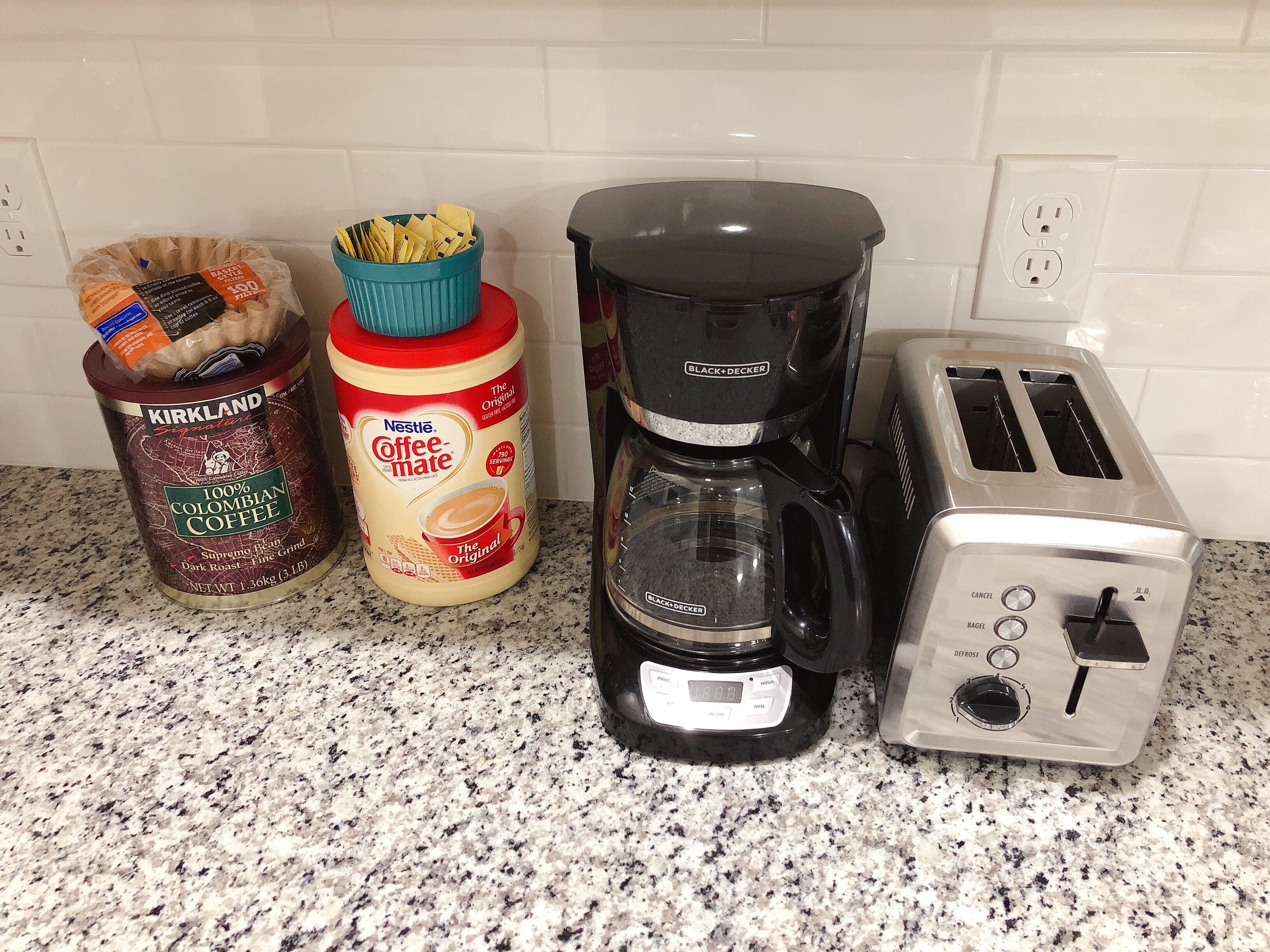 Un plan de travail de cuisine avec une machine à café, un grille-pain, un récipient de crème à café et un paquet de dosettes de café.