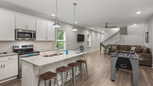 A modern kitchen with a central island, bar stools, a foosball table, and a staircase leading to a living area with a sofa. - Luxurious 8 bd/6ba with ROOFTOP! Price is the total! (Nashville)