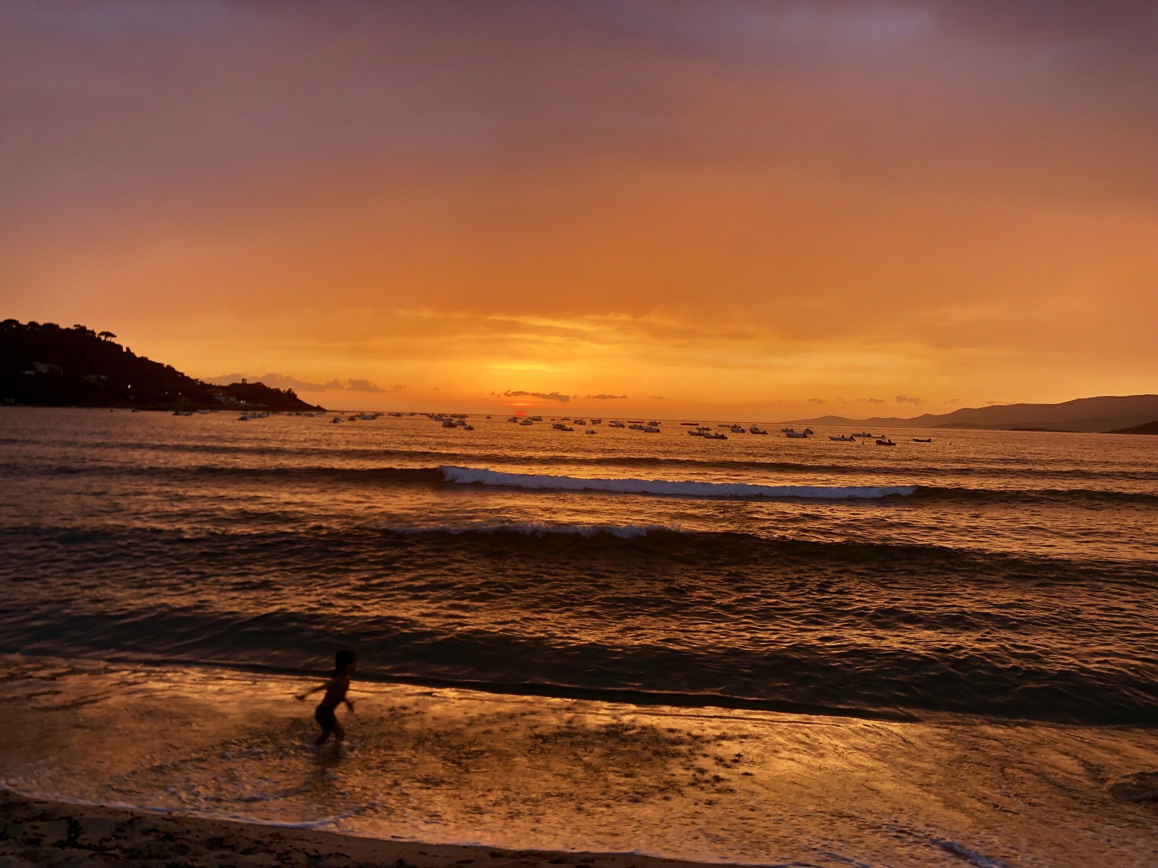 A sunset over a beach with boats and a child playing in the water.