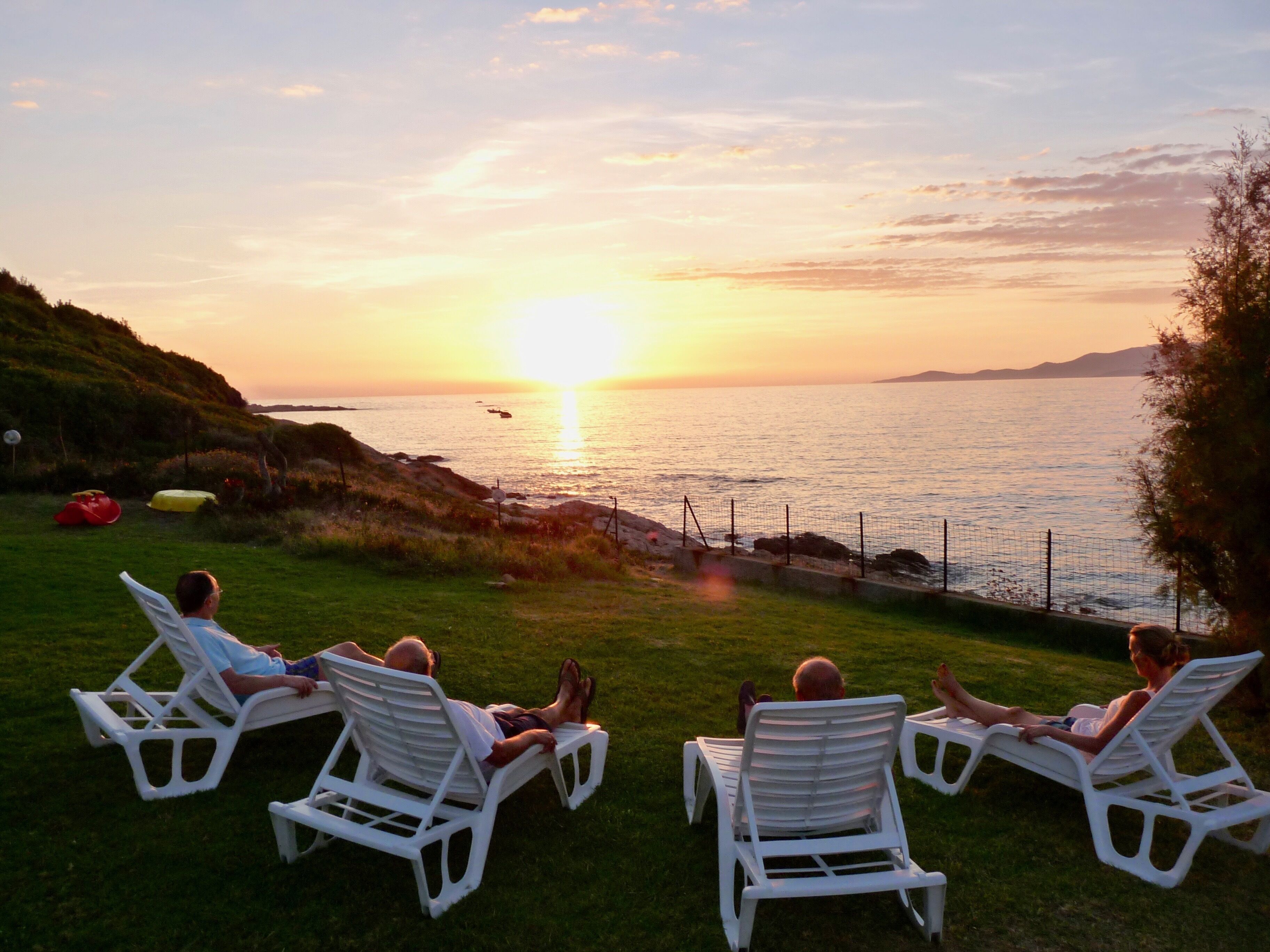 A sunset view from a grassy area with white lounge chairs facing the sea.
