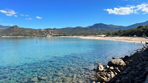 A coastal beach with clear water, rocky shoreline, and distant mountains.