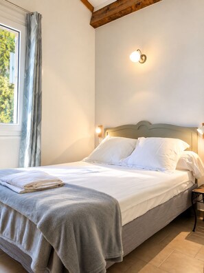 A neatly made bed with a grey blanket and white pillows, a bedside table with a lamp, and a window with curtains.