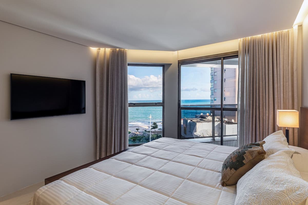 A hotel room with a large bed, a TV mounted on the wall, and a view of the ocean through a large window.