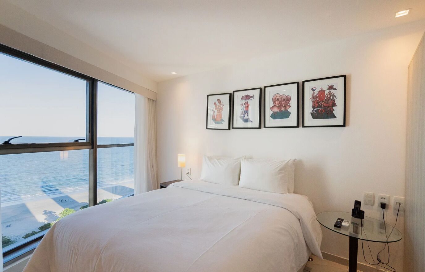 A hotel room with a large bed, a glass table with electronic devices, and a view of the water outside.