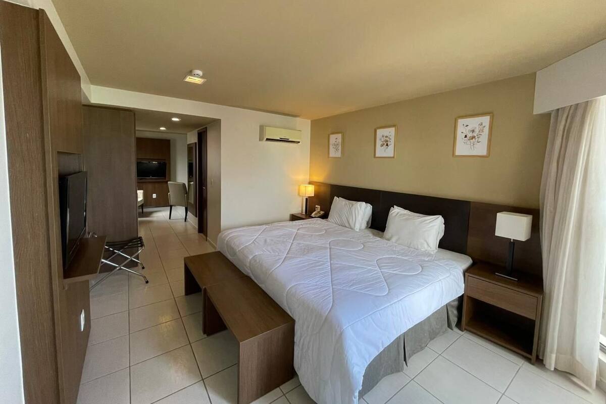 A hotel room with a bed, bedside tables, a TV, a bench, and a desk.