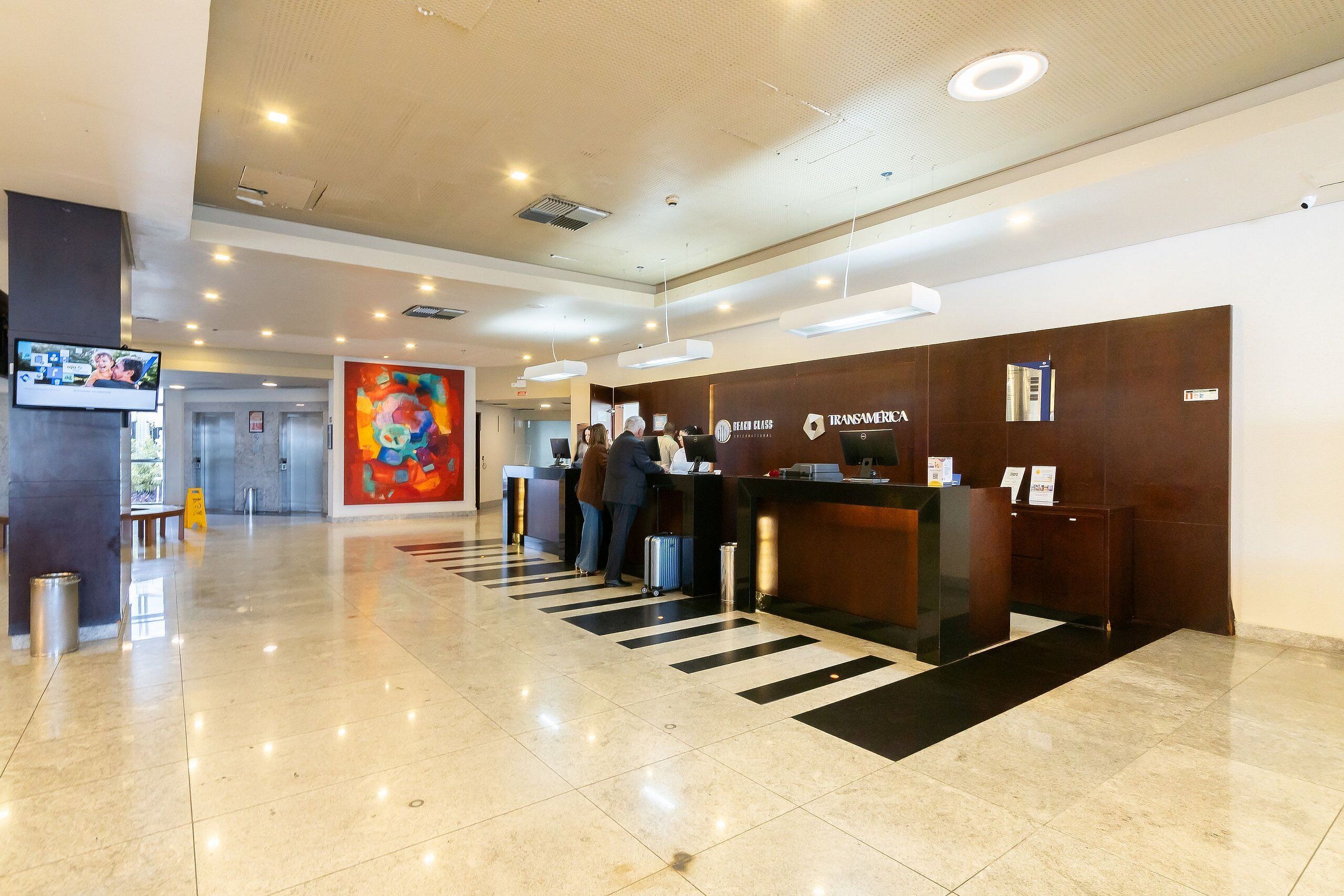 Zona de recepción de un hotel con diseño moderno, que incluye un mostrador de recepción, una pantalla digital y un cuadro abstracto de gran tamaño.