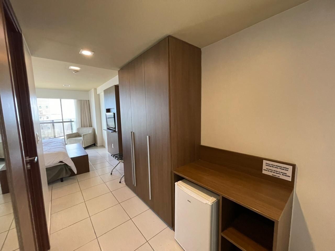 A compact hotel room with a kitchenette, a bed, a small seating area, and a view of the outside.