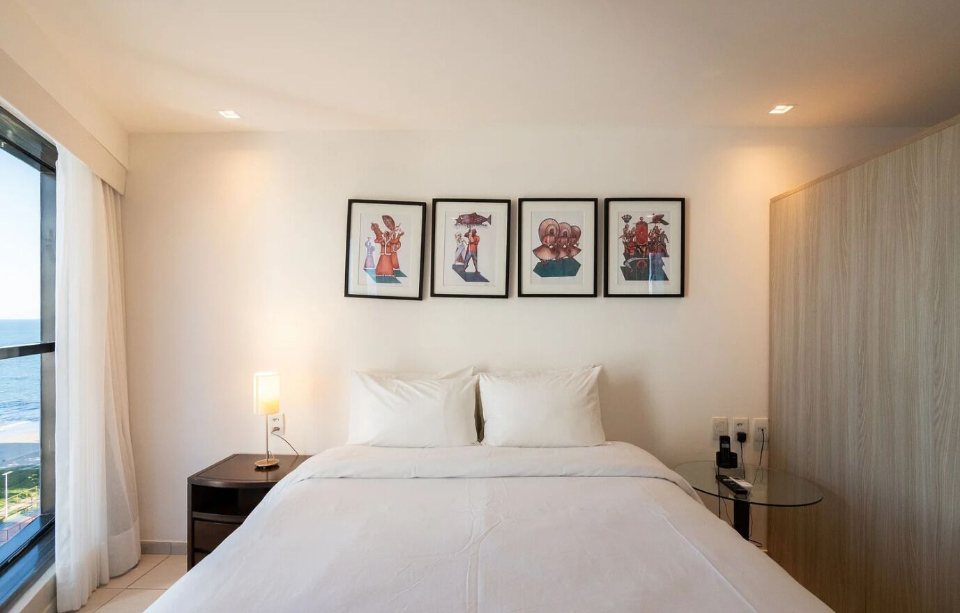 A hotel room with a bed, a bedside table with a lamp, a small table with a phone and charger, and a window with a view of the sea.