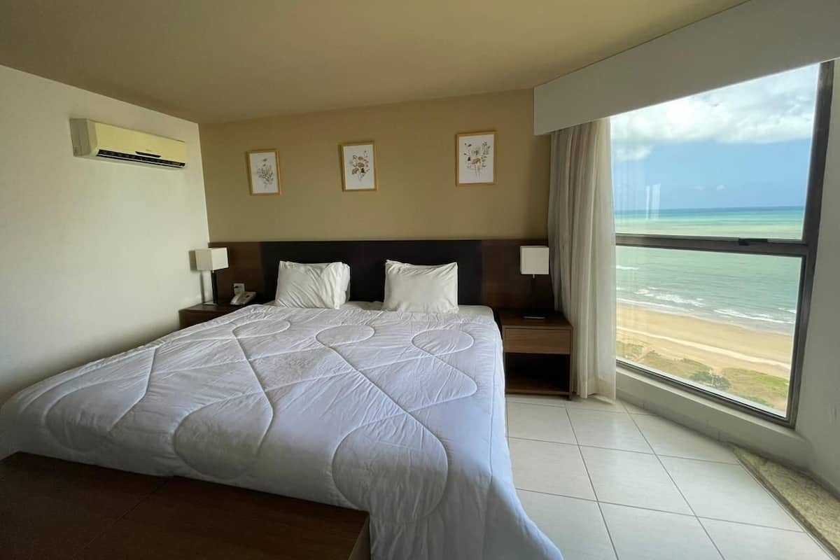 A hotel room with a large bed, a nightstand, a bedside lamp, a wall-mounted air conditioner, and a large window with a beach view.