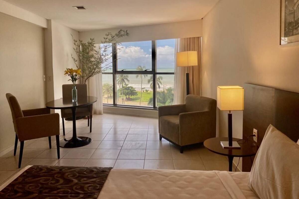 A hotel room with a bed, a desk with a chair, a small table with a lamp, and a window with a view of the beach.