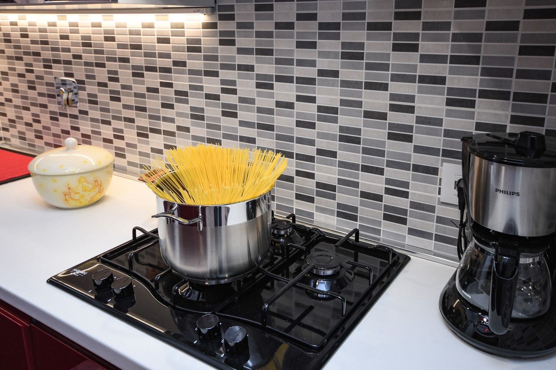 Una cocina con estufa a gas, una olla de espagueti, una cafetera y un cuenco decorativo.