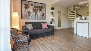 Ein modernes Wohnzimmer mit grauem Sofa, einem Metallstuhl, einer Weltkarte als Kunstwerk, einem weißen Bücherregal und einer Küchenzeile mit weißem Kühlschrank und einer Kaffeemaschine.