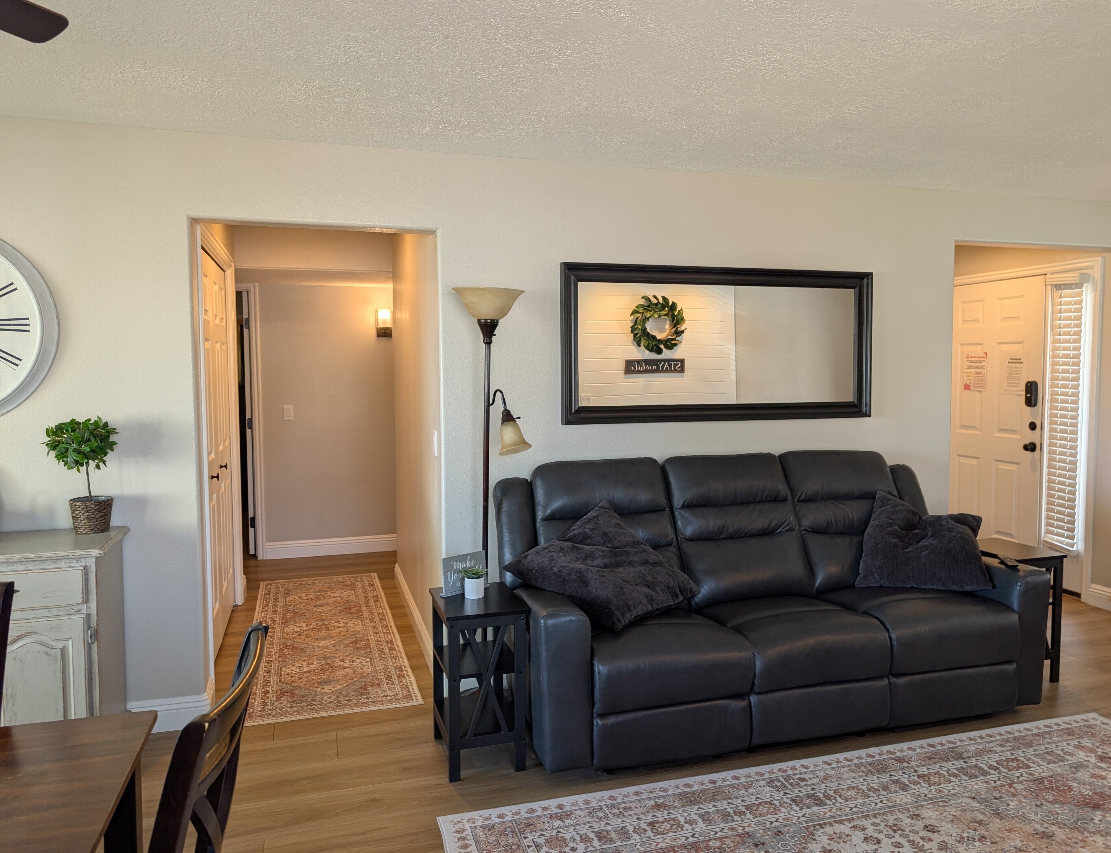 A living room with a black leather sofa, a wall clock, a floor lamp, a rug, and a door with a sign.