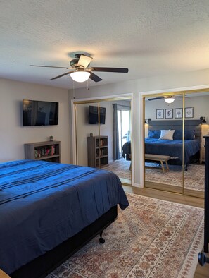A bedroom with a bed, a ceiling fan, a TV, a bookshelf, and a large mirror.