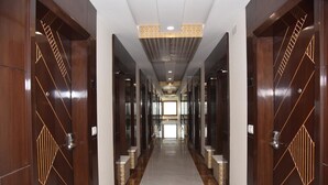 Um corredor comprido e estreito com portas de madeira adornadas com padrões geométricos, que dá passagem a um quarto claro no final.