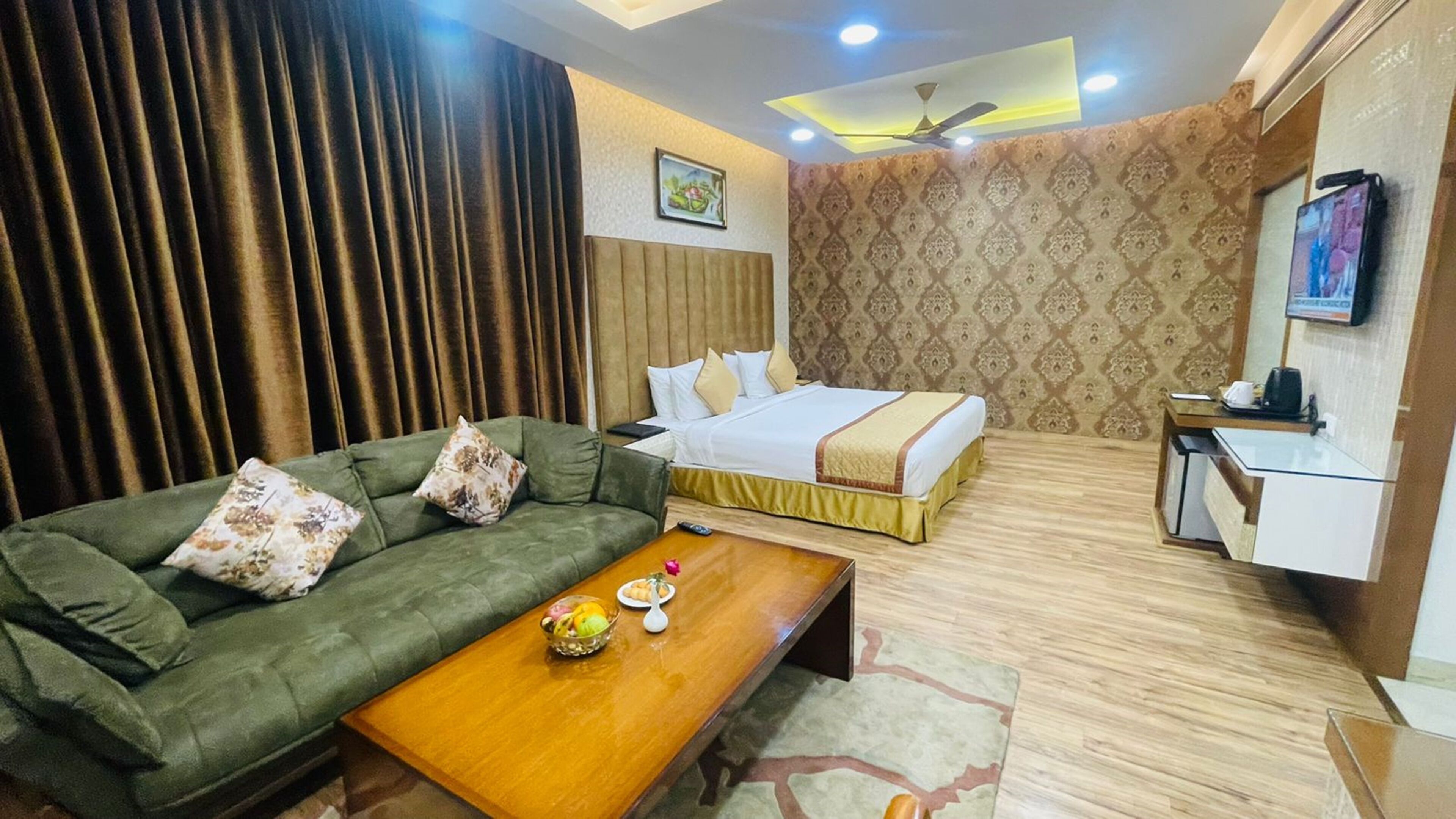 Quarto de hotel com sofá verde, cama com roupa de cama branca, mesa de centro de madeira com frutas e televisão na parede.