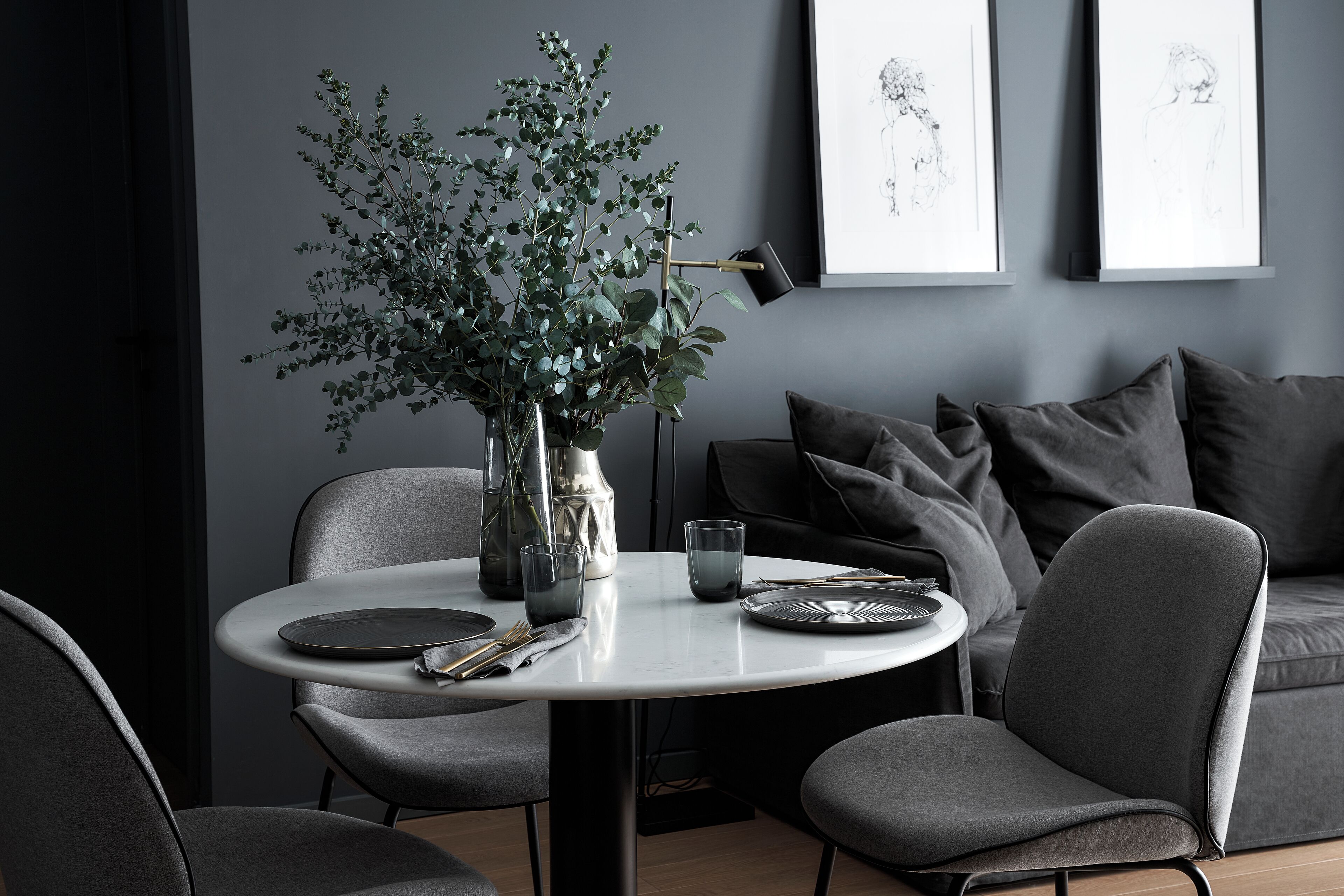 A modern dining area with a round table, grey chairs, a vase with green plants, and framed artwork on the wall.