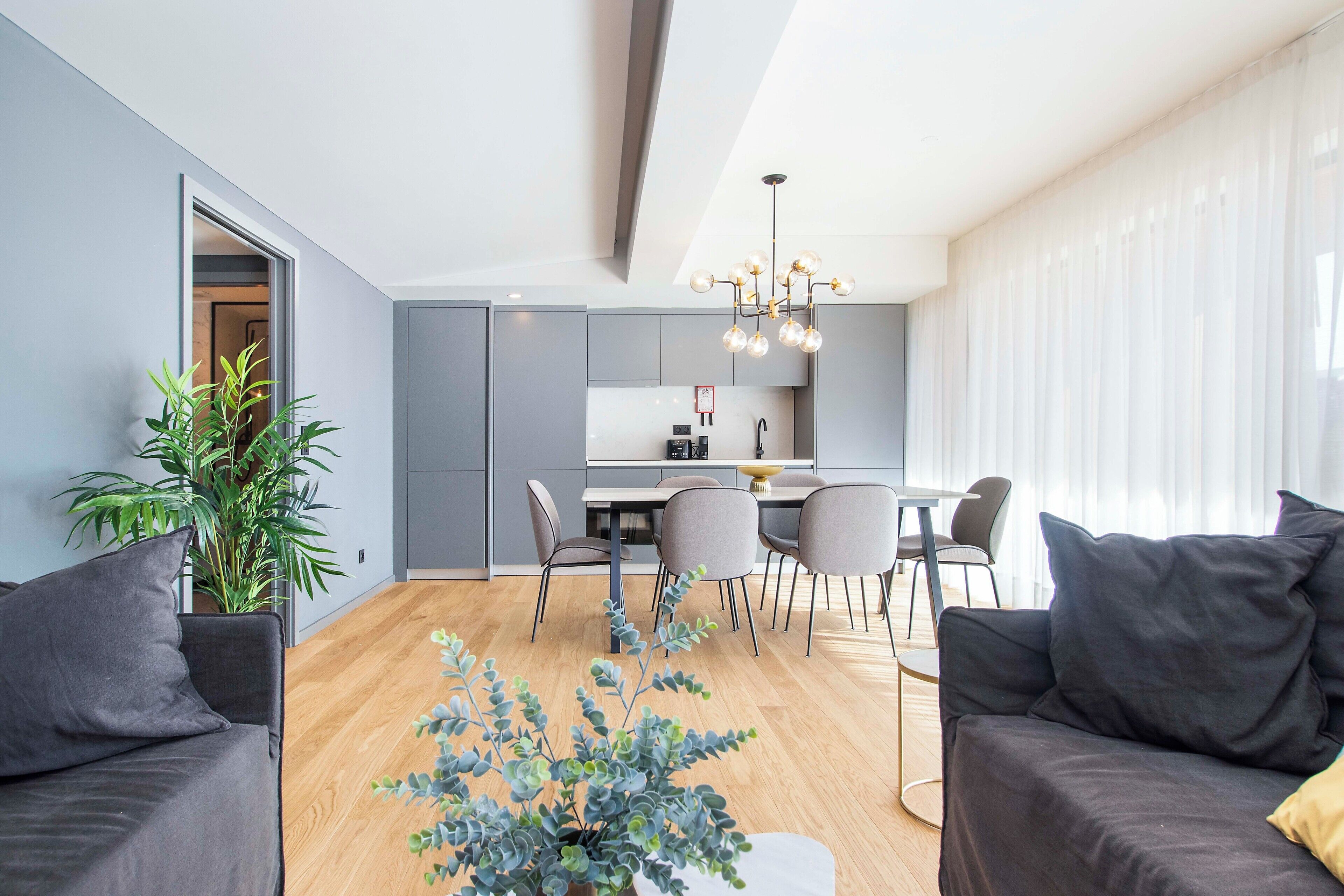 A modern living room with a grey sofa, a dining area with a table and chairs, and a kitchen in the background.
