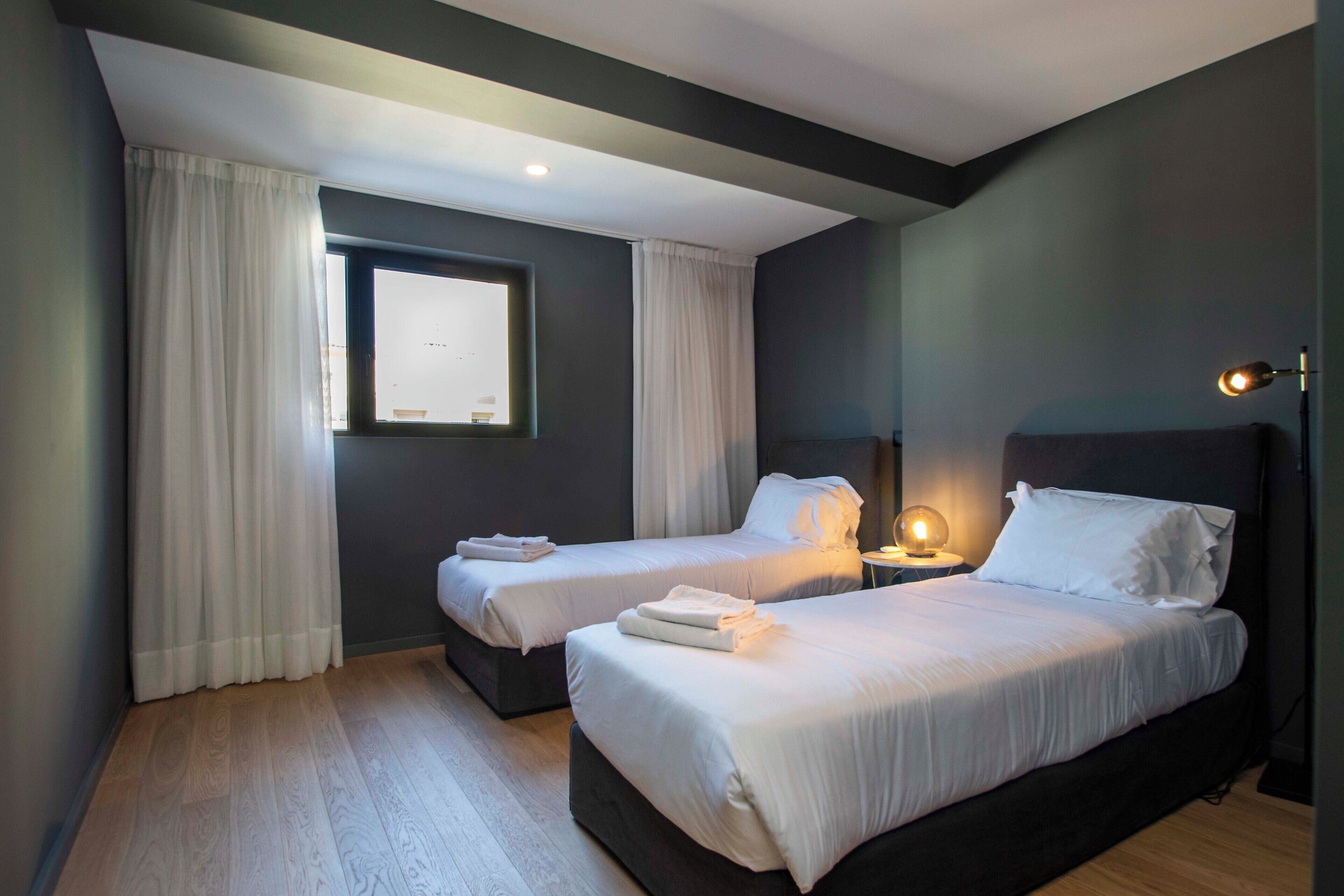 A hotel room with two beds, a window with curtains, a bedside lamp, and a view of the outside.