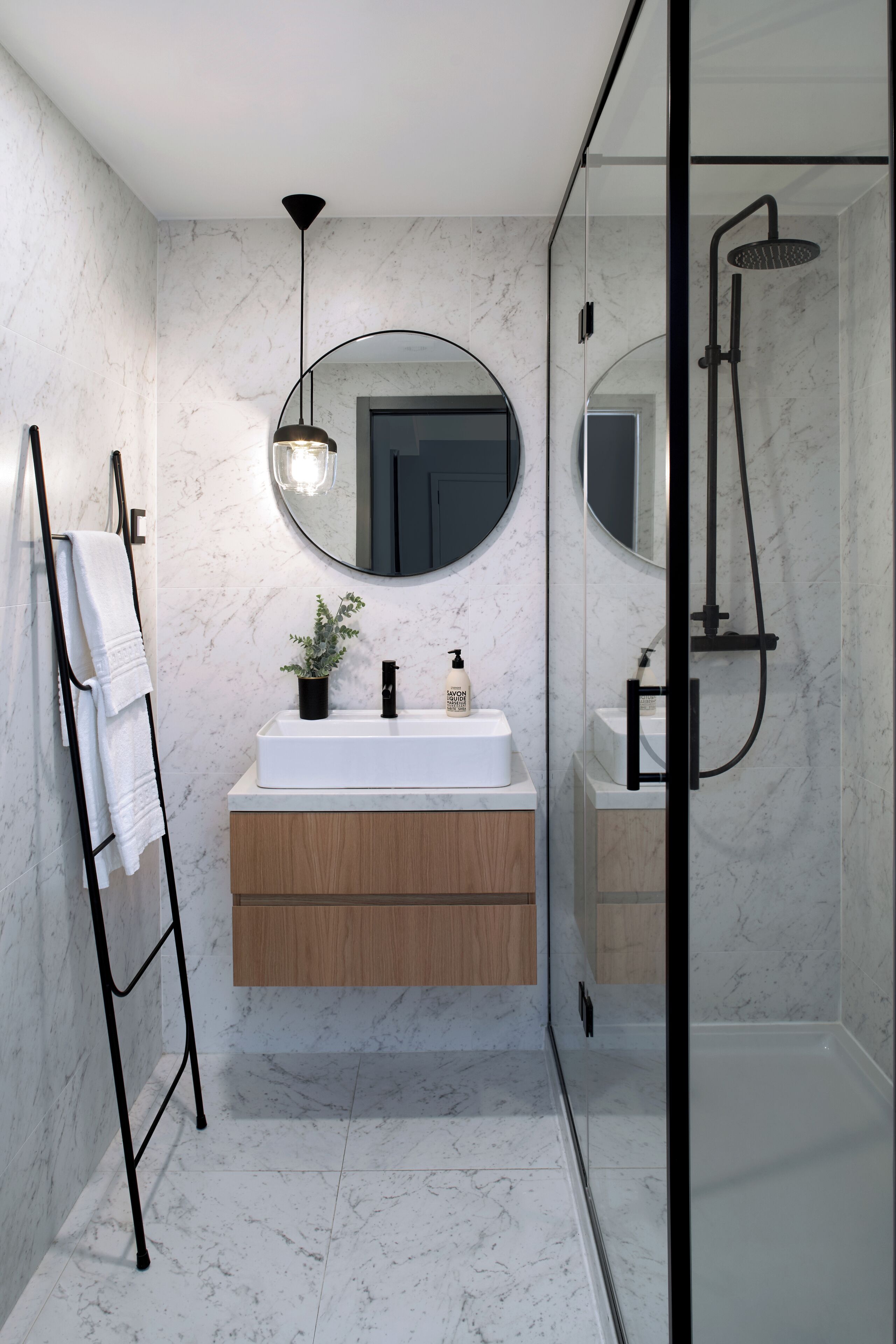 Een moderne badkamer met een marmeren vloer, een houten wastafelmeubel, een ronde spiegel en een douchecabine met glazen wanden.