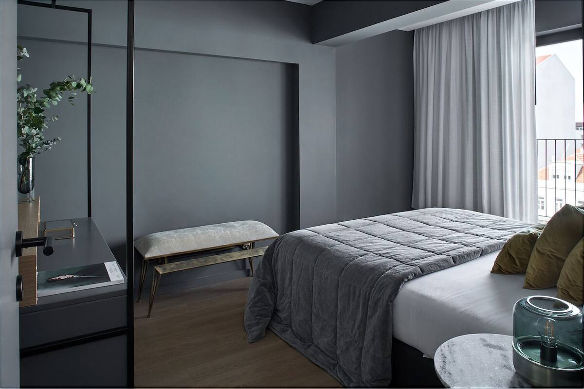 Un dormitorio moderno con cama, banca, mesita de noche y un florero con flores.