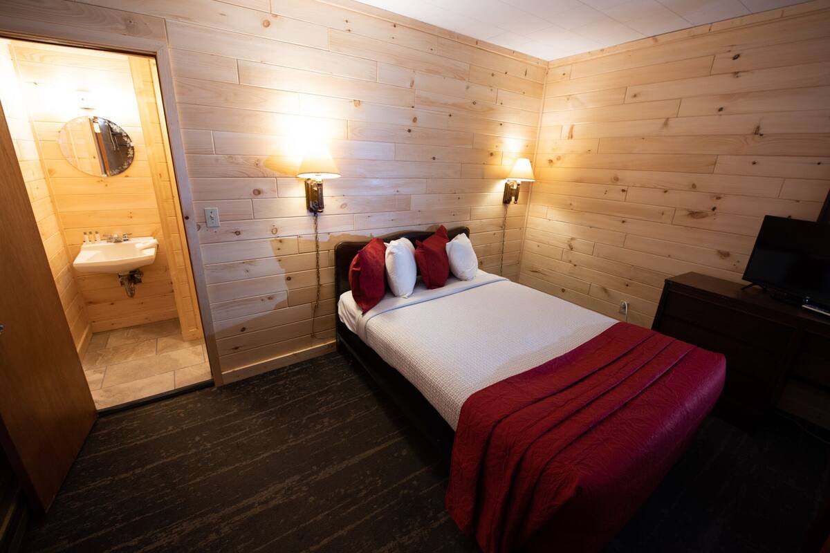 A wooden-paneled room with a bed, two wall-mounted lamps, a TV, and a bathroom with a sink and mirror.