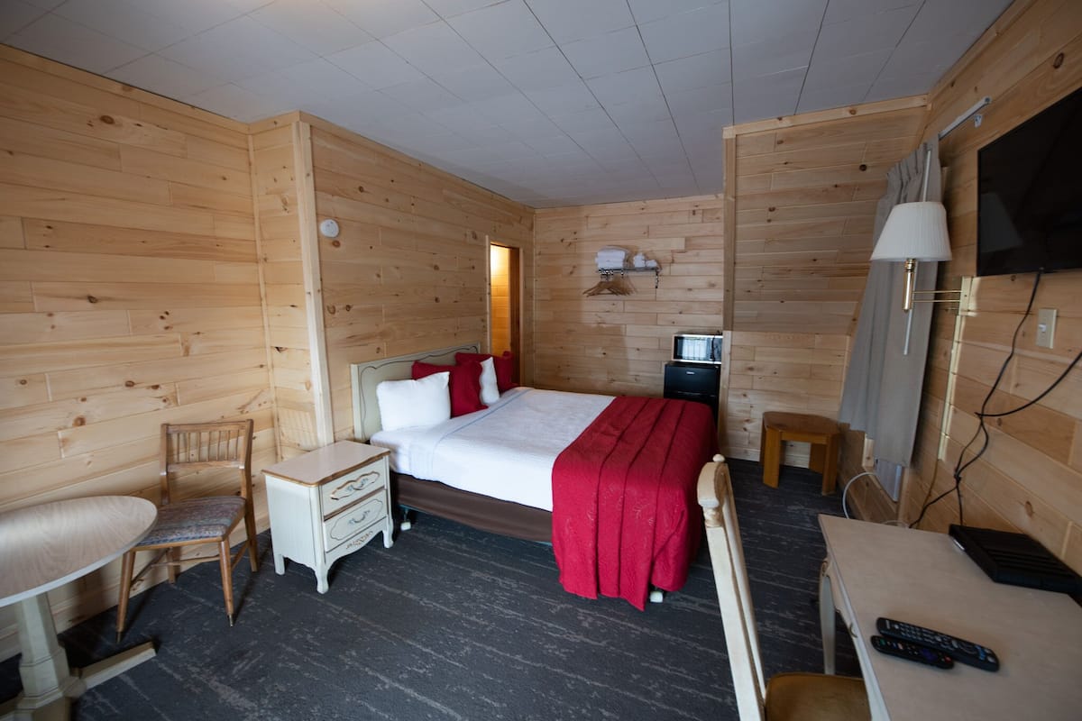 A wooden-paneled room with a bed, a desk, a chair, a TV, and a lamp.