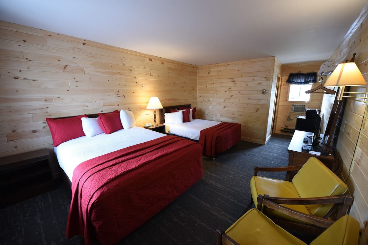A hotel room with wooden walls, two beds with red and white bedding, a yellow chair, a desk with a lamp, and an air conditioning unit.