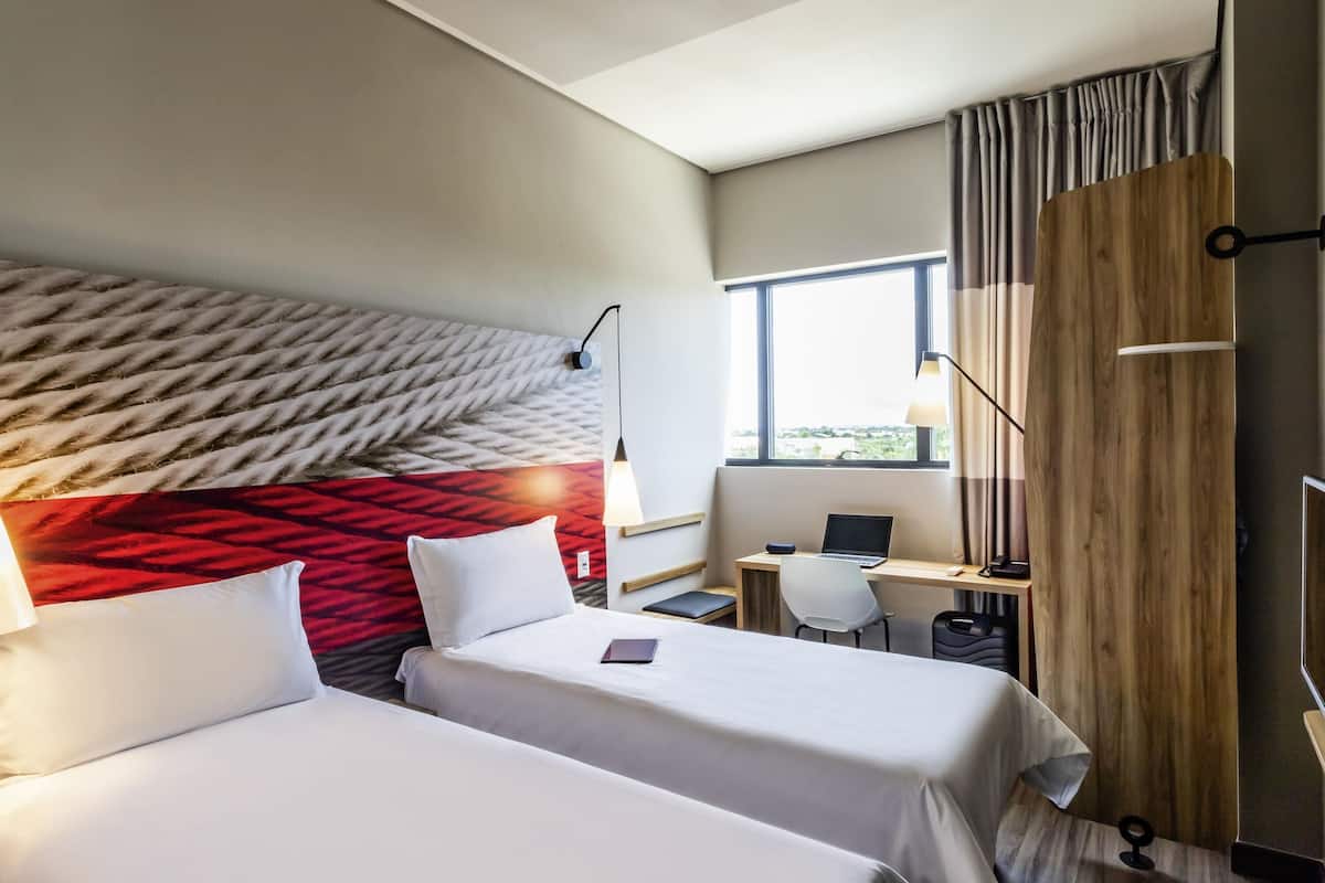 A hotel room with two beds, a desk with a laptop, a chair, and a large window.