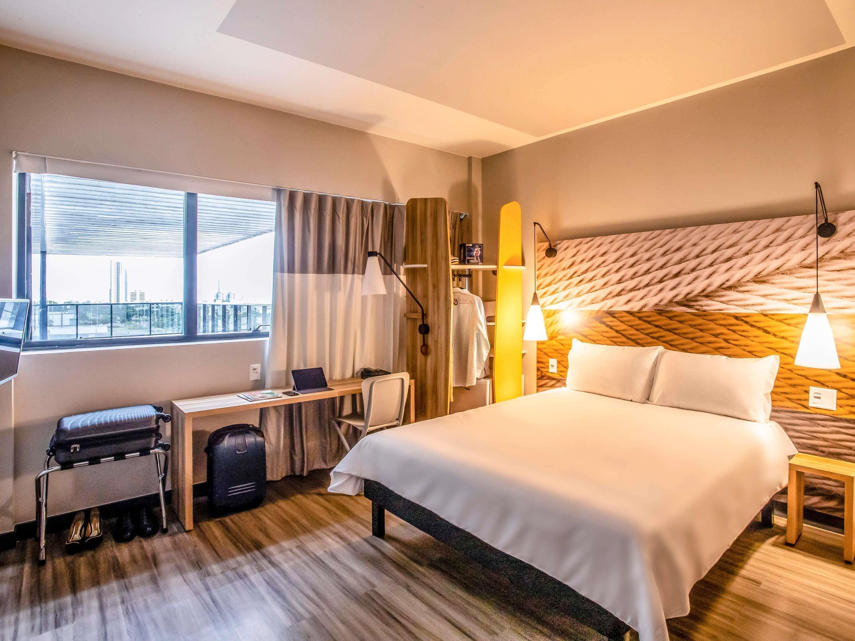 Ein Hotelzimmer mit Bett, Schreibtisch, Stuhl, Koffer und Blick auf die Stadt.