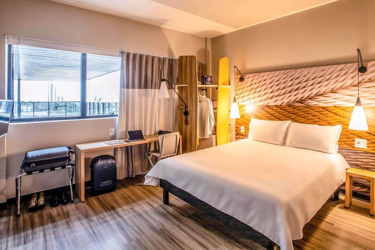 A hotel room with a bed, desk, chair, suitcase, and a view of the city.