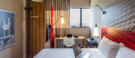 Habitación de hotel con cama, escritorio con una computadora portátil, una silla, ventana y pared con un cuadro.