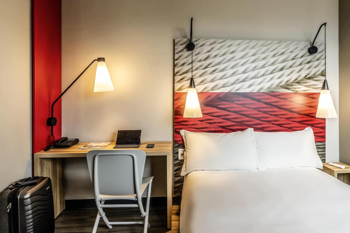 A hotel room with a bed, desk, chair, and a suitcase.
