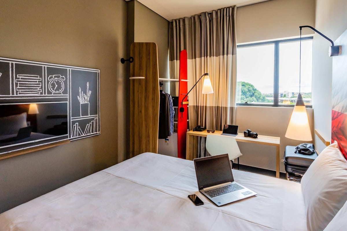 A hotel room with a bed, a desk with a laptop, a desk lamp, and a window with a view of greenery.