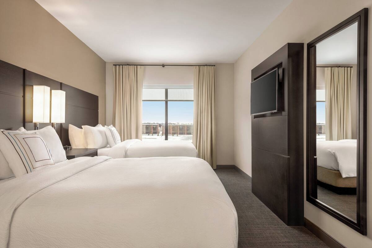 Ein Hotelzimmer mit zwei Betten, einem großen Spiegel, einem Flachbildfernseher und Blick auf die Stadt durch das Fenster.
