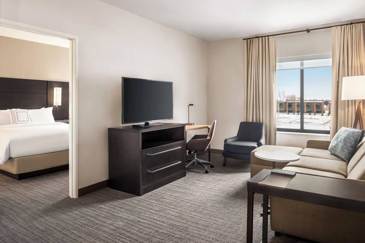 Ein Hotelzimmer mit Bett, Schreibtisch mit Stuhl, Sofa, Fernseher und einem Fenster mit Vorhängen.