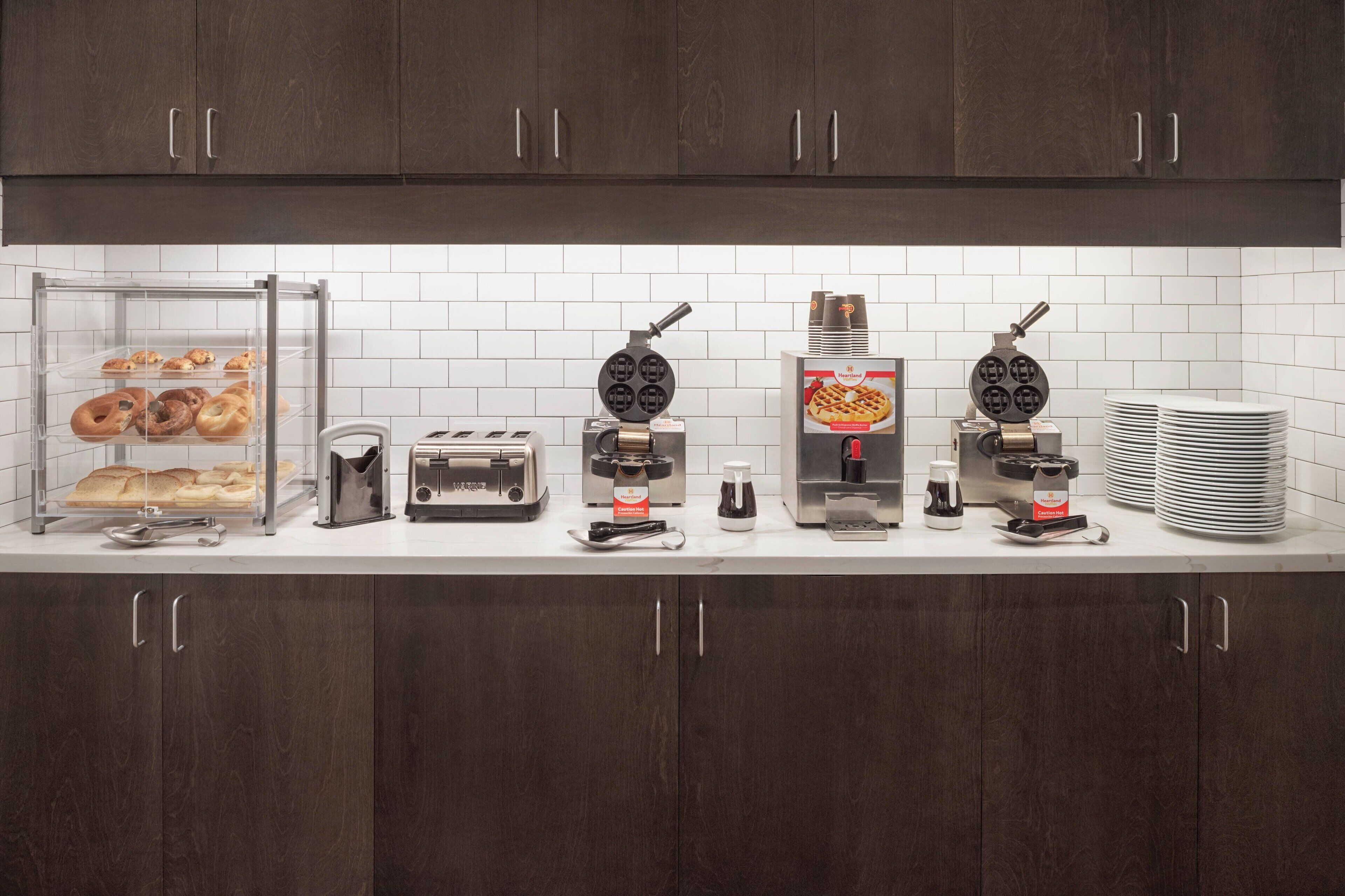 A kitchen with dark wooden cabinets, white tiled backsplash, and various appliances including a waffle maker, toaster, and coffee maker.