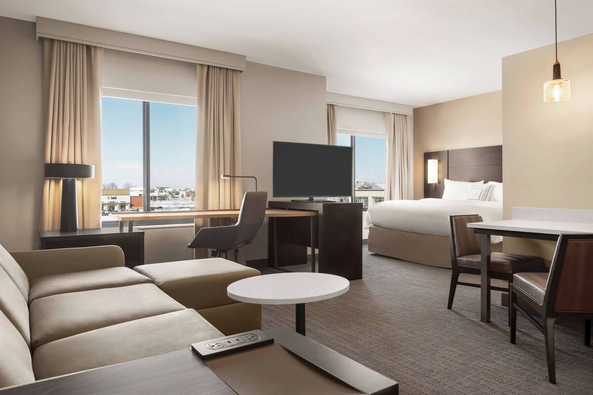 Ein Hotelzimmer mit einem großen Bett, einem Schreibtisch mit Flachbildfernseher, einer Couch, einem kleinen runden Tisch und einem Sessel.