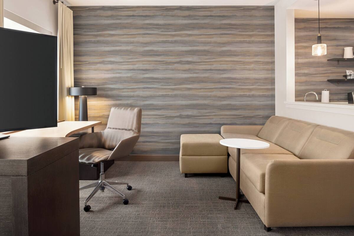 Ein modernes Hotelzimmer mit Schreibtisch, gepolstertem Sessel, Sofa und Wand mit Muster.