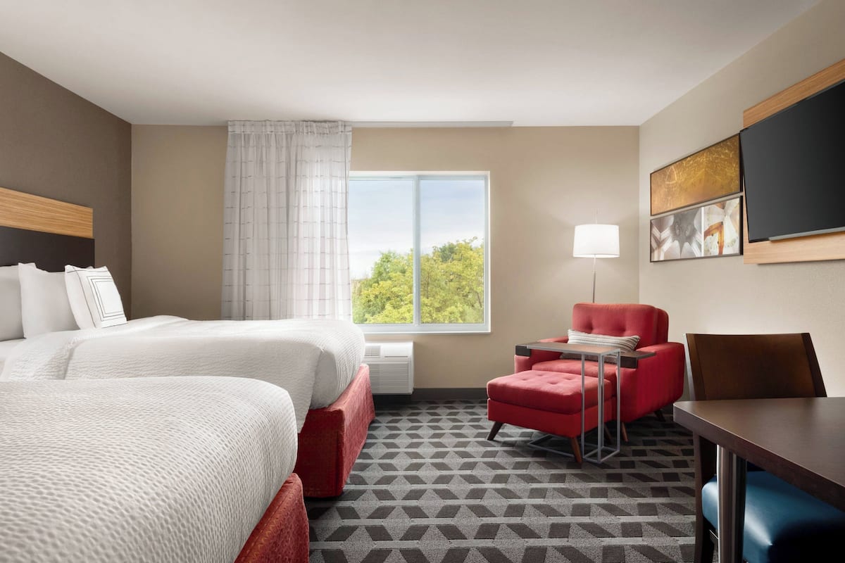 A hotel room with two beds, a red armchair, a table, and a large window with a view of trees.