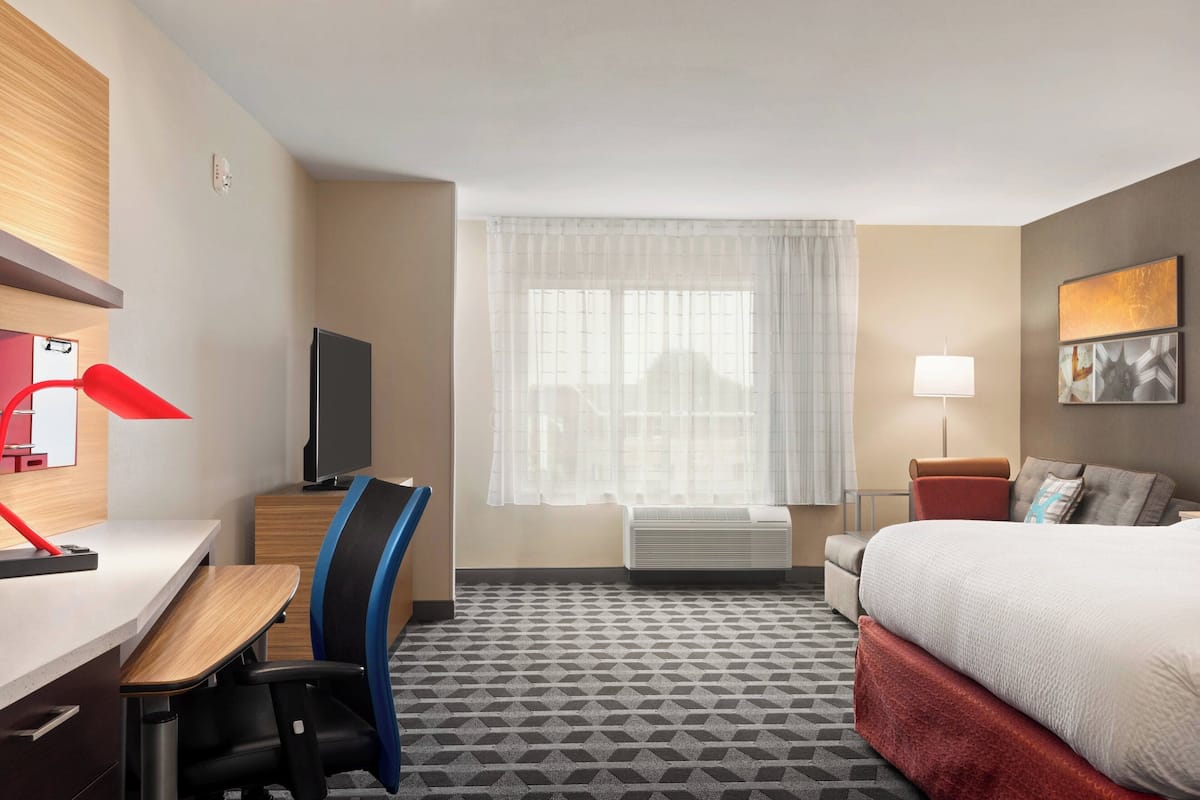 A hotel room with a bed, desk, chair, television, window with curtains, and a lamp.