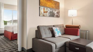 A modern hotel room with a grey sofa, a side table, a lamp, and a bed with a red bedspread. - TownePlace Suites by Marriott Janesville (Janesville)