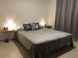Un lit bien fait, recouvert d’une couverture et agrémenté d’oreillers, deux tables de chevet avec des lampes, et un rideau gris à la fenêtre.